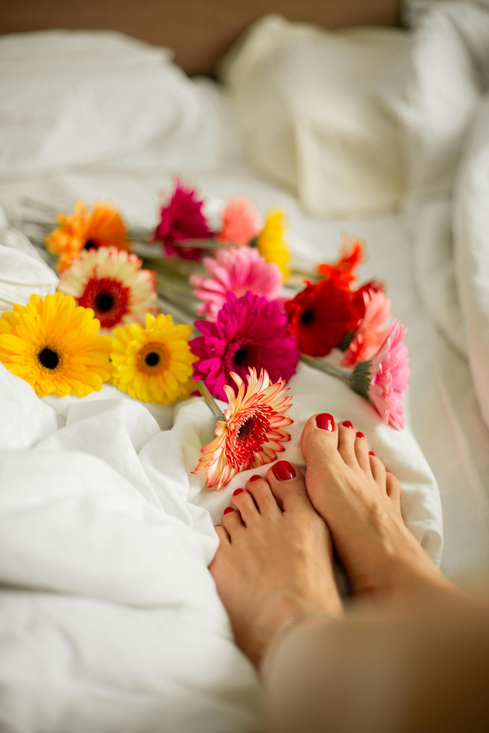 A woman's feet rest on white bedding adorned with colorful gerbera flowers, creating a delicate and relaxing scene.